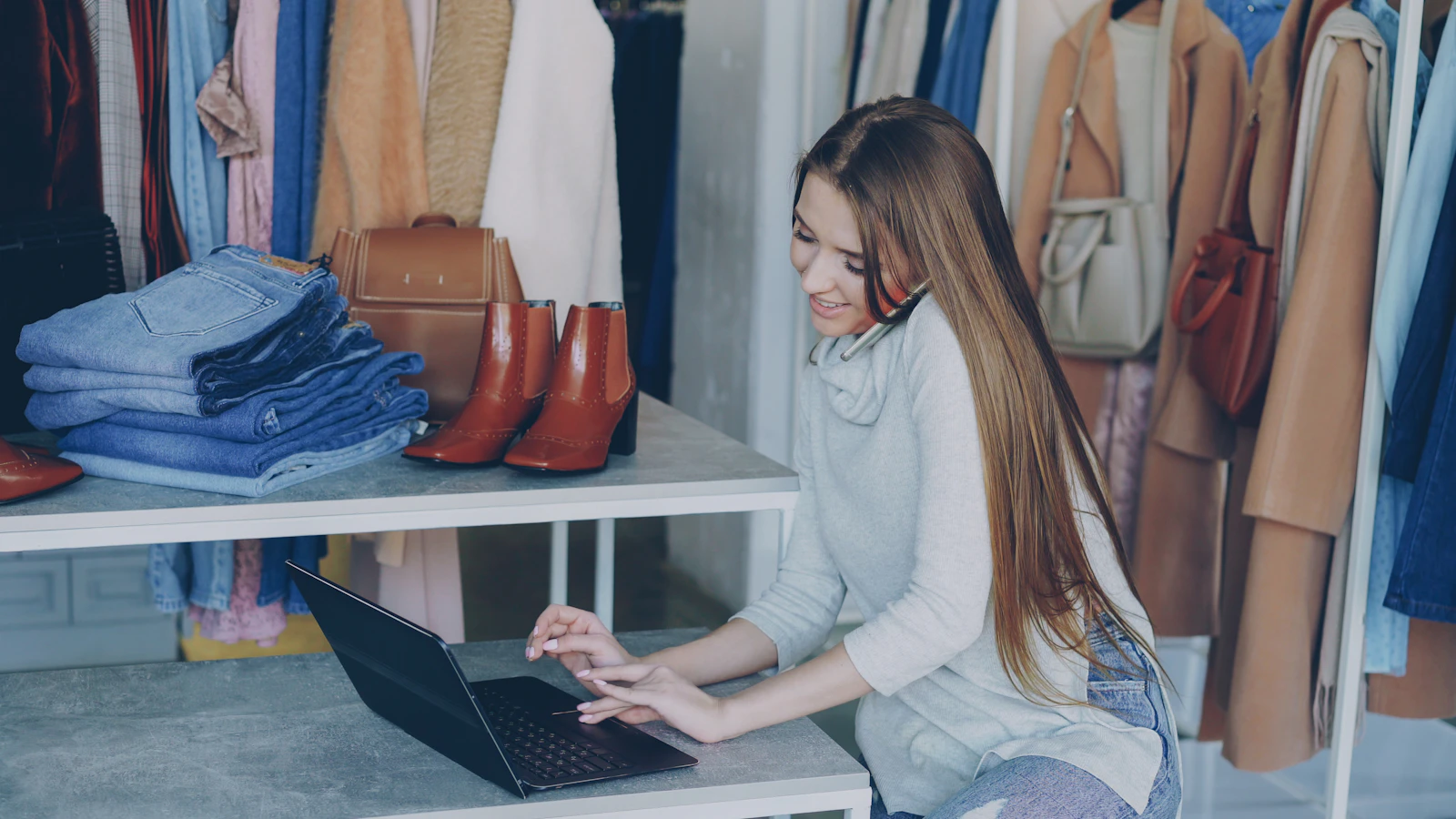 Chủ shop thời trang vừa nghe điện thoại vừa gõ laptop giữa kệ quần áo